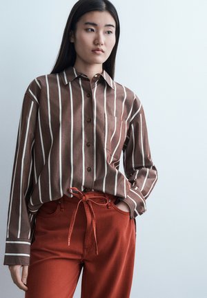 Femme aux longs cheveux noirs portant une chemise à rayures verticales marron et blanches, rentrée dans un pantalon à cordon de serrage couleur rouille, les mains dans les poches.