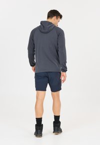 Man draagt een donkergrijze capuchontrui, marineblauwe short, zwarte sokken en zwarte sneakers, staand met de rug naar voren op een effen witte achtergrond.