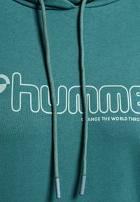 Teal hoodie med dragsko, med en vit logotyp som säger "humme" och frasen "FÖRÄNDRA VÄRLDEN GENOM." Mjuk, strukturerad tyg.