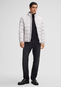 Gepolsterte, hellgraue Daunenjacke mit hohem Kragen, Reißverschluss und Seitentaschen, kombiniert mit schwarzen Jeans und schwarzen Schuhen.