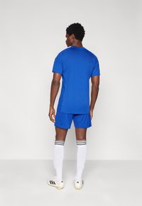 Άνδρας που στέκεται με τη πλάτη προς το φακό, φορώντας μπλε ποδοσφαιρική στολή, λευκές κάλτσες μέχρι το γόνατο με μαύρες ρίγες και λευκά παπούτσια ποδοσφαίρου με μαύρες ρίγες.