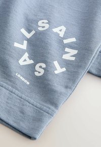 Tissu bleu clair avec un texte blanc disposé en cercle indiquant "SAINT SALTS" et un texte plus petit "LONDON" en dessous.