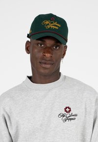 Donkergroene pet met gele en rode borduursel aan de voorkant, gedragen met een lichtgrijze sweatshirt met een logo op de borst.