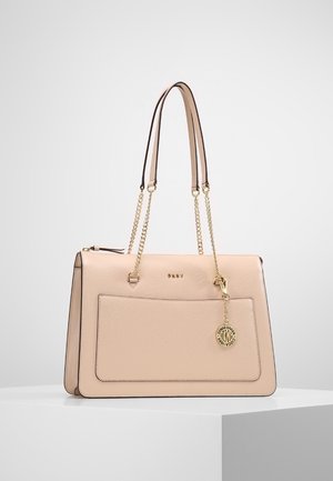 Beżowa skórzana torebka DKNY z podwójnymi złotymi łańcuszkowymi paskami, przednią kieszenią i okrągłą złotą zawieszką z logo projektanta, wisząca na białej powierzchni.