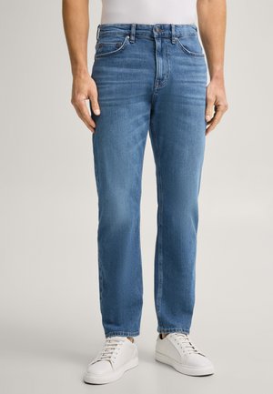 Person wearing medium blue straight-leg jeans, white sneakers, and a white top, standing against a plain light background.
