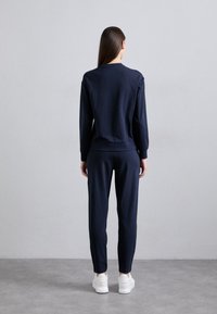 Sieviete ar gariem brūniem matiem stāv ar muguru vērsta, ģērbusies tumši zilos sporta tērpos un baltos sporta apavos uz pelēka grīdas un sienas fona.