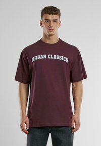 Bordo kokvilnas T-krekls ar īsām piedurknēm, apaļu kaklu un "URBAN CLASSICS" uzrakstu kontrastējoši gaiši zili krāsā krūšu daļā.