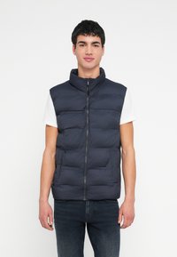 Granatowa kamizelka puchowa z wysokim kołnierzem i przednim zamkiem błyskawicznym. Posiada pikowane sekcje i lekki, wodoodporny materiał. Noszona na białej koszuli.