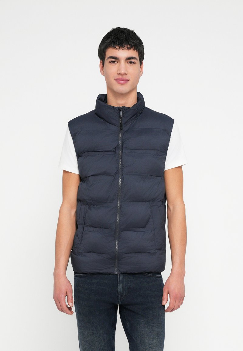 Granatowa kamizelka puchowa z wysokim kołnierzem i przednim zamkiem błyskawicznym. Posiada pikowane sekcje i lekki, wodoodporny materiał. Noszona na białej koszuli.