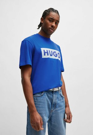 Blue cotton t-shirt with a white rectangular "HUGO" logo, round neckline, and short sleeves, paired with light blue denim jeans.