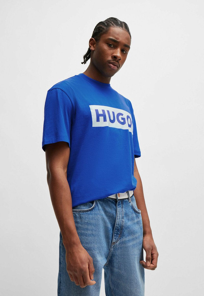 Blå bomull T-skjorte med en hvit rektangulær "HUGO"-logo, rund hals og korte ermer, sammen med lyseblå denimbukser.