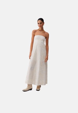 Donna con un abito bianco lungo fino al pavimento senza spalline, ricamato con sottili motivi floreali, stivali beige, collana d'oro e orecchini, in piedi davanti a uno sfondo semplice.