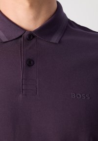 Temno vijolična polo majica z dvema gumboma, teksturirana tkanina in subtilen logotip vtisnjen na prsih. Klasična oblika ovratnika.