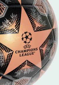 Balón de fútbol de la UEFA Champions League en negro y naranja que presenta paneles con estrellas y patrones de hojas, con el logo centrado debajo. Textura suave y brillante.