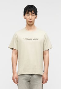 Μπεζ βαμβακερό T-shirt με στρογγυλή λαιμόκοψη και κοντά μανίκια. Διαθέτει ανάγλυφο λογότυπο "MICHAEL KORS" στο στήθος. Συνδυάζεται με ανοιχτό γκρι παντελόνια.