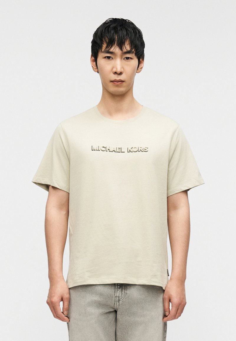 Μπεζ βαμβακερό T-shirt με στρογγυλή λαιμόκοψη και κοντά μανίκια. Διαθέτει ανάγλυφο λογότυπο "MICHAEL KORS" στο στήθος. Συνδυάζεται με ανοιχτό γκρι παντελόνια.