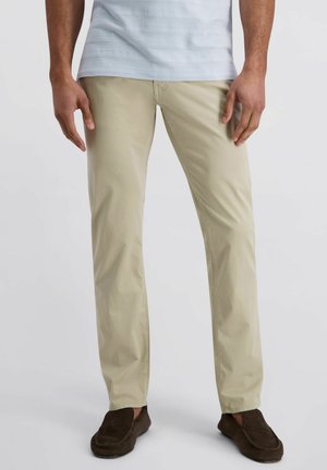 Homme portant un pantalon beige coupe droite, une chemise rayée bleu clair et des mocassins marron foncé, debout devant un fond clair.
