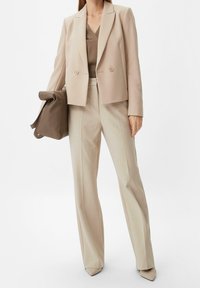 Blazer beige à double boutonnage porté sur un chemisier taupe, accompagné d'un pantalon beige clair sur mesure et d'un sac à main marron. Escarpins beige pointus.