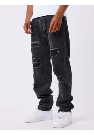 Personne portant un jean noir usé et des baskets blanches, debout devant un fond clair uni.
