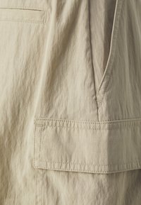 Beige tkanina s texturovaným povrchem, zdobená náprstní kapsou a viditelným švem podél okrajů. Neformální design s čistými liniemi.