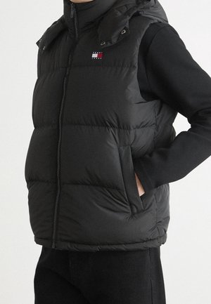 Person trägt schwarze Steppweste mit kleinem Logo auf der Brust, Reißverschluss vorne und Händen in den Seitentaschen über einem schwarzen Langarmshirt.
