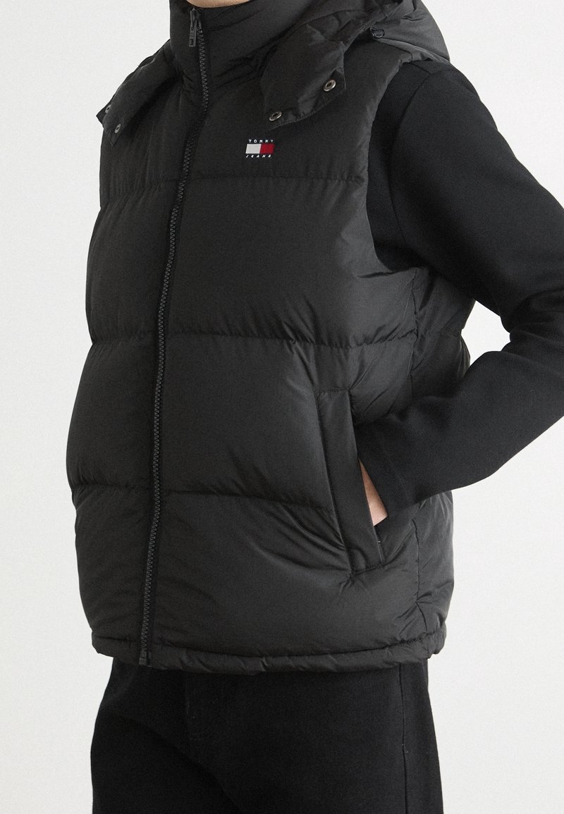 Personne portant une veste matelassée noire avec un petit logo sur la poitrine, fermeture éclair à l'avant et les mains dans les poches latérales par-dessus un haut noir à manches longues.