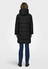 Doudoune noire avec capuche, dotée d'un design matelassé et de manches longues. Portée par-dessus un jean bleu et des chaussures noires.