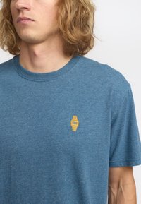 Camiseta de algodón azul con un pequeño diseño de reloj bordado en amarillo en el lado izquierdo del pecho. Cuello redondo y mangas cortas. Textura suave.