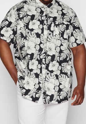 Zwart overhemd met korte mouwen en knopen, bedrukt met een witte tropische bloemenprint, gecombineerd met een witte broek, zichtbaar van schouders tot midden bovenbeen.