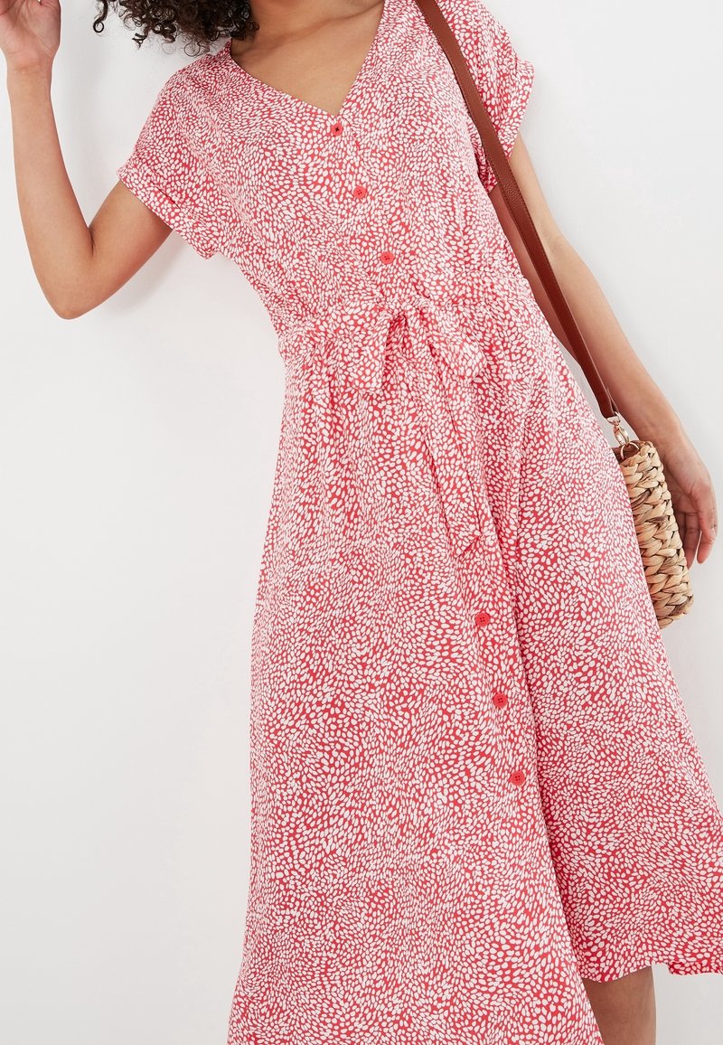Femme portant une robe boutonnée à manches courtes rouge et blanche avec une ceinture à la taille et portant un sac bandoulière tressé.