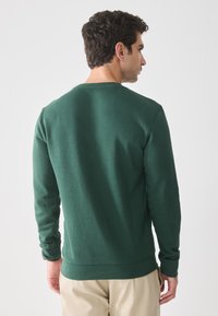Uomo con capelli ricci scuri, indossa una camicia a maniche lunghe verde scuro e pantaloni beige, in piedi di spalle su uno sfondo bianco.