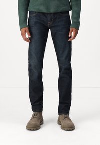 Jeans in denim blu scuro con vestibilità slim, caratterizzati da cuciture arancioni e tasche frontali, abbinati a stivali alla caviglia in suede beige.