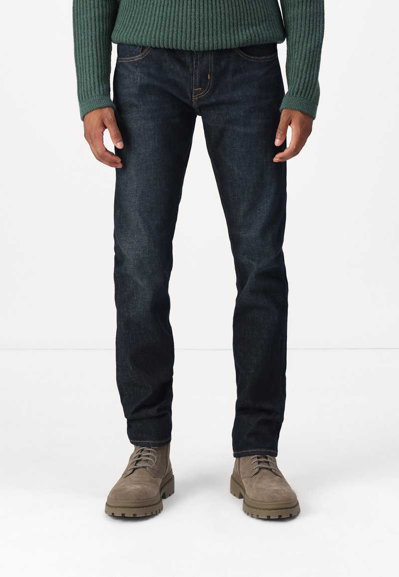 Jeans in denim blu scuro con vestibilità slim, caratterizzati da cuciture arancioni e tasche frontali, abbinati a stivali alla caviglia in suede beige.
