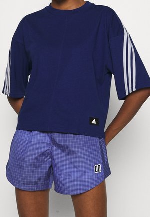 Osoba nosząca granatowy t-shirt Adidas z białymi paskami na rękawach oraz fioletowe szorty w kratę z wzorem "00" na lewej nodze.