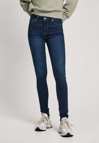 Jeans ajustados de denim oscuro, con un ligero brillo, bolsillos frontales y costuras en contraste, combinados con zapatillas de color claro.