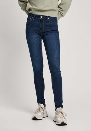 Jeans aderenti in denim scuro, con una leggera lucentezza, tasche frontali e cuciture a contrasto, abbinati a scarpe da ginnastica chiare.