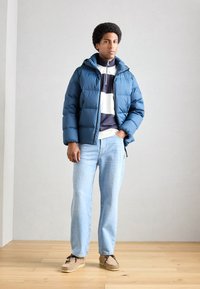 Pardoseală albastră cu glugă, cu un design matlasat. Asortată cu un pulover cu dungi, blugi deschiși la culoare și pantofi bej.