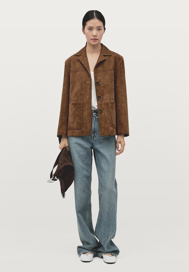 Brązowa zamszowa kurtka z kołnierzem z wycięciem, dwiema przednimi kieszeniami i czarnymi guzikami, zestawiona z jasnoniebieskimi, szerokimi dżinsami i białymi butami.