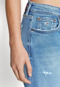 High-waisted blue denim jeans with a distressed patch, featuring copper-tone hardware and a small branded tag near the pocket.