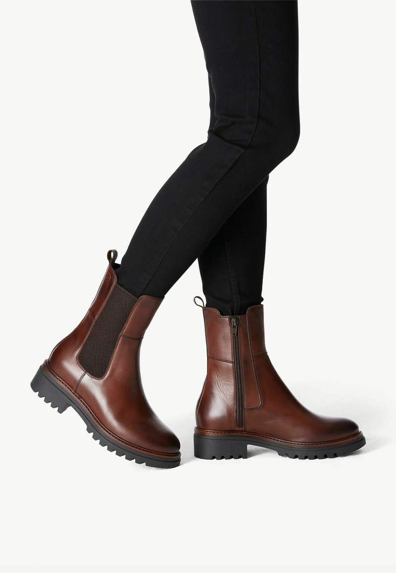 Person som bär mörkbruna läderankelstövlar med sidizipper och elastisk panel, i kombination med svarta åtsittande byxor.