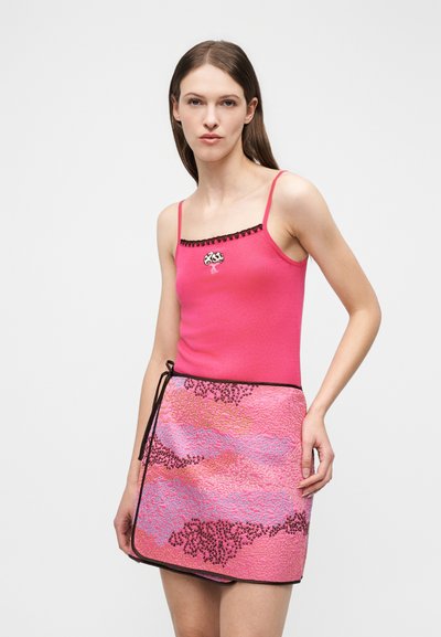 Γυναίκα με ροζ καλοκαιρινό τοπ με μαύρο περίγραμμα και κεντημένο δέντρο, συνδυασμένο με φούστα τύπου σαρί σε ροζ, μαύρο και μπλε σχέδιο.