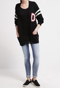 Svart cardigan med vita ränder och ett rött bokstav "D", med knappar, fickor och en avslappnad passform, i kombination med ljusblå smala jeans.