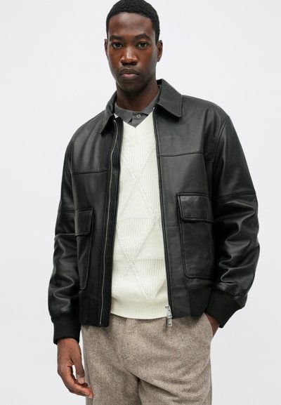 Giubbotto bomber in pelle nera con zip frontale, due grandi tasche e polsini a coste. Indossato sopra un maglione cremoso a trama e pantaloni a fantasia.