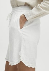 Shorts en lin blancs avec une taille élastique, détail de cordon de serrage, poches latérales et bord froncé au bas. Texture lisse, coupe décontractée.