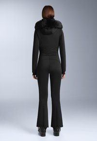 Czarny kombinezon z kołnierzem z sztucznego futra, charakteryzujący się dopasowanym krojem, teksturowanym pasem i rozszerzanymi nogawkami z zamkami na mankietach.