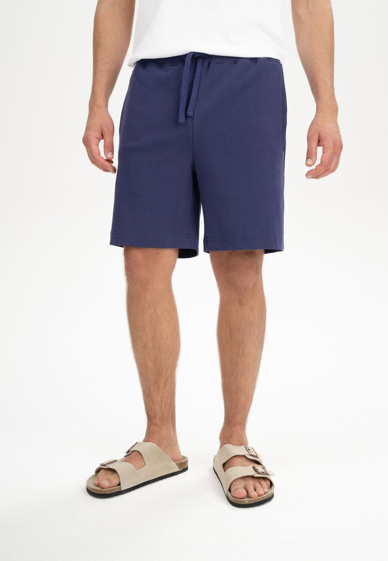 Marineblaue Baumwollshorts mit elastischem Bund und Kordelzug, ausgestattet mit Seitentaschen und einem lockeren Schnitt, kombiniert mit beigen Sandalen.