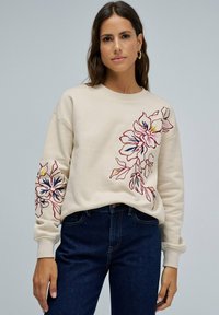 Mujer con sudadera beige decorada con bordados florales en rosa, amarillo y marino en el pecho y la manga izquierda, combinada con jeans azul oscuro.