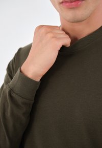 Olivgrüner Sweatshirt mit geripptem Kragen und Bündchen, aus weichem Stoff hergestellt. Eine Nahaufnahme zeigt eine Hand, die den Ausschnitt anpasst und die Textur hervorhebt.