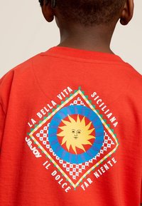 Rode katoenen sweatshirt met een kleurrijk zonontwerp in een diamantvorm, omringd door tekst en geometrische patronen in diverse kleuren.