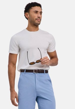 Mann mit weißem kurzärmligen Hemd und hellblauer Hose, hält eine schwarze Sonnenbrille in der rechten Hand, steht vor einem schlichten Hintergrund.